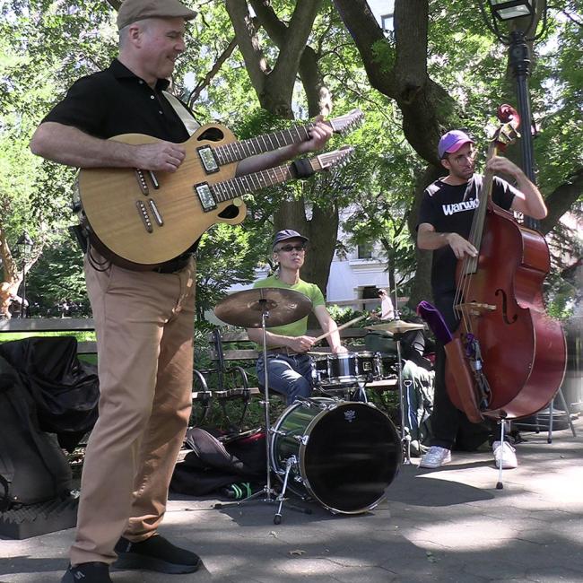 Scott plays the double-necked guitar in a park with a drummer and a standing bass player. 