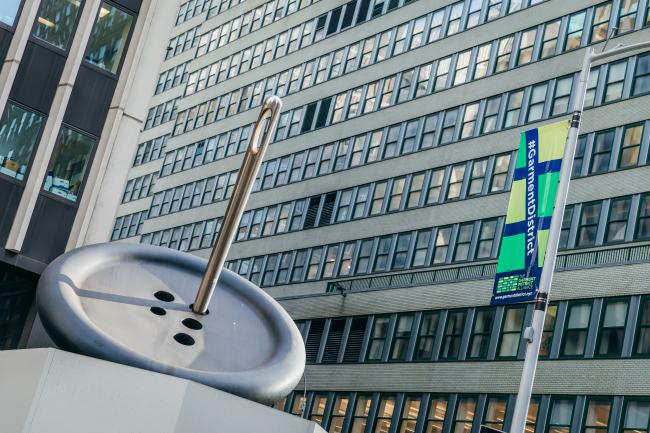 A view of the Button & Needle sculpture with a Garment District banner and an office building in background