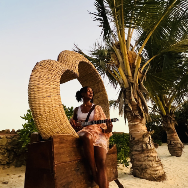 A young woman sits on an ornate chair on the sand playing guitar with palm trees behind her. 
